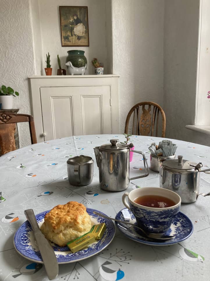 Ein altmodischer Raum, auf einemm runden Tisch mit Tischdecke steht ein Gedeck mit Tee und einem Scone, Teekanne, Milchkännchen aus Edeelstahl daneben